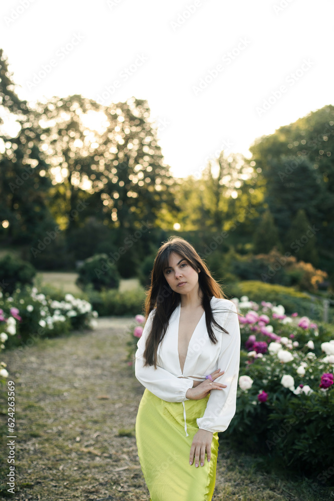beautiful young brunette woman in white blouse and light green satin skirt posing while standing in the garden. caucasian girl posing near peonies in the park. beautiful young brunette woman in white blouse and light green satin skirt posing while standing in the garden. caucasian girl posing near peonies in the park.