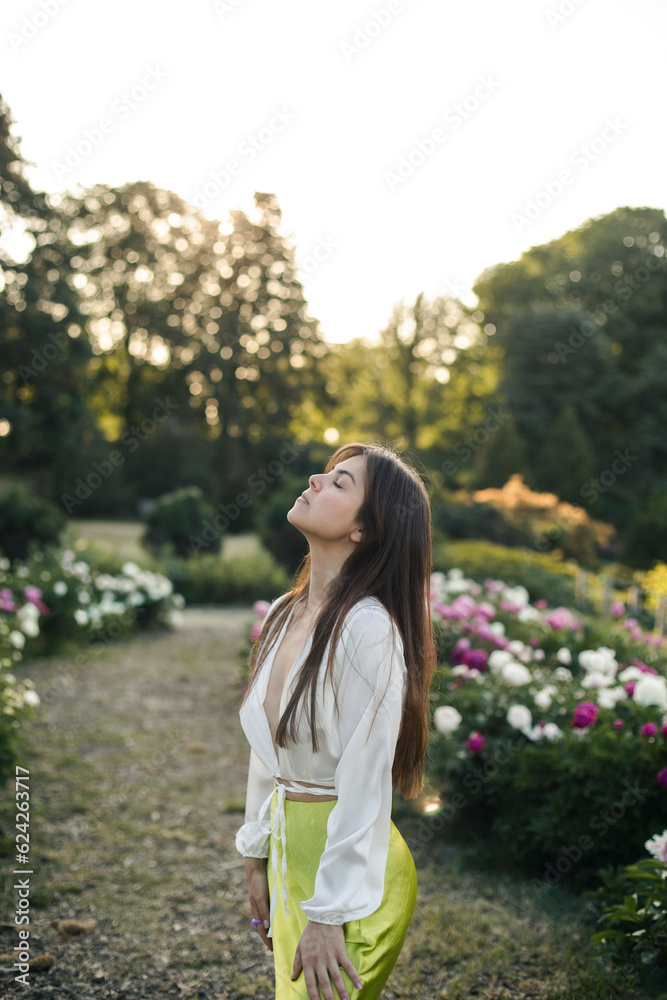 a beautiful young brunette woman in a white blouse and light green satin skirt enjoys the sunrise in the garden. caucasian girl posing near peonies in the park. a beautiful young brunette woman in a white blouse and light green satin skirt enjoys the sunrise in the garden. caucasian girl posing near peonies in the park.