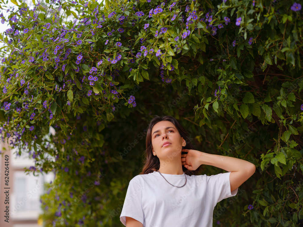 Beautiful young woman with dark curly hair and perfect skin wearing white tshirt posing near blooming flowers in garden. Nude make up. Allergy season, antihistamines. Aroma oils natural perfumes Beautiful young woman with dark curly hair and perfect skin wearing white tshirt posing near blooming flowers in garden. Nude make up. Allergy season, antihistamines. Aroma oils natural perfumes