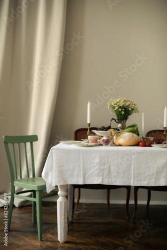 vintage white table with white tablecloth in a room with beige walls and antique armchairs. decorated table with appetizers, bread, vegetables, berries and a vase of daisies.	