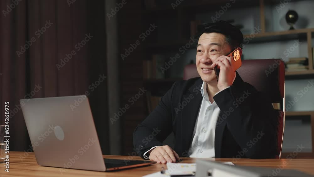 Angle view of self-confident asian real estate agent sitting in modern office with personal computer and talking on cellphone with customer. Cheerful man in stylish black suit making offers for sale. Angle view of self-confident asian real estate agent sitting in modern office with personal computer and talking on cellphone with customer. Cheerful man in stylish black suit making offers for sale.