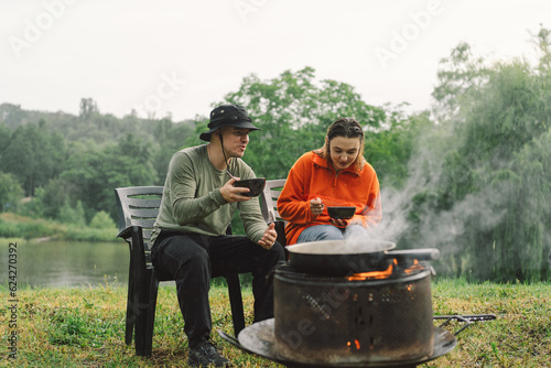 A man and a woman enjoy the food by the fire. Hiking camp with a bonfire in the forest. Tourist on recreation outside. Campsite lifestyle