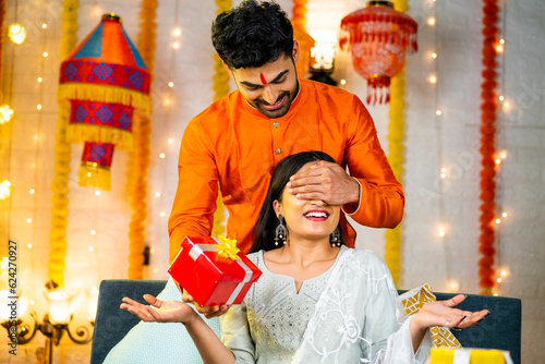 Fotografie Happy indian man by closing eyes giving surprise gift to wife during festival celebration at home - concept of relationship bonding, togetherness and raksha Bandhan