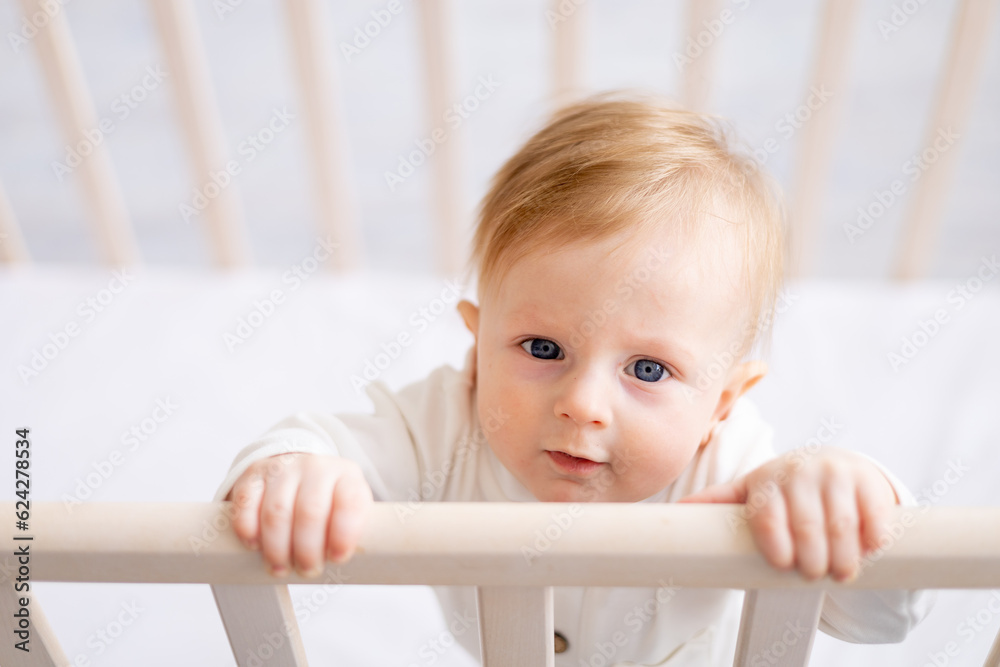 close-up of a smiling baby 6 months old blond boy in a crib in a bright bedroom in a white cotton bodysuit, portrait, concept of children's goods close-up of a smiling baby 6 months old blond boy in a crib in a bright bedroom in a white cotton bodysuit, portrait, concept of children's goods