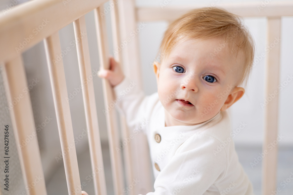 close-up of a smiling baby 6 months old blond boy in a crib in a bright bedroom in a white cotton bodysuit, portrait, concept of children's goods