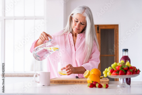Mature woman pouring detox water in glass at home photo
