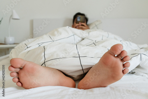 Young man with using smart phone lying barefoot on bed at home