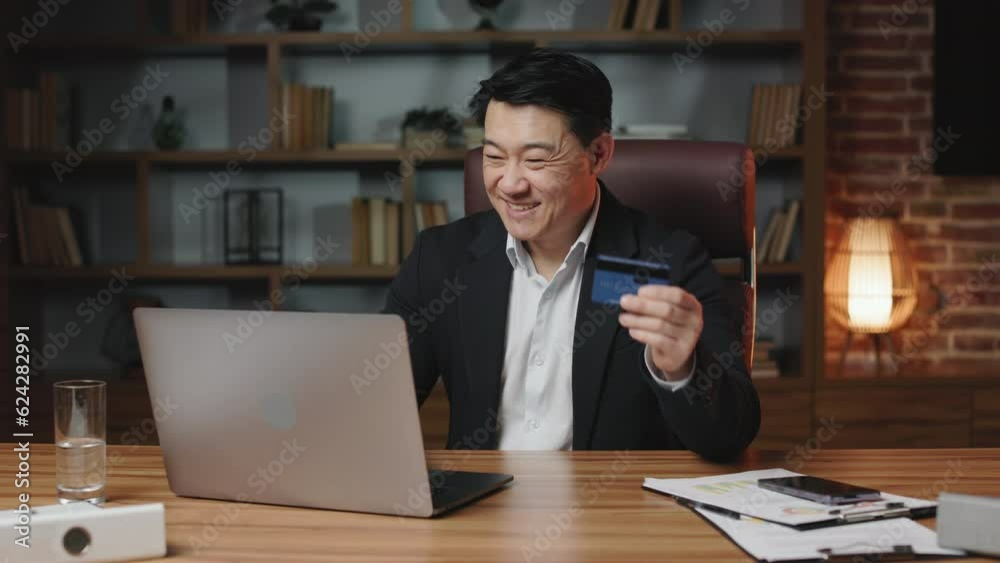Cheerful asian employee having access to unlimited credit card and doing online shopping in comfy office. Smiling man dressed in black suit making payment on laptop via internet banking. Cheerful asian employee having access to unlimited credit card and doing online shopping in comfy office. Smiling man dressed in black suit making payment on laptop via internet banking.