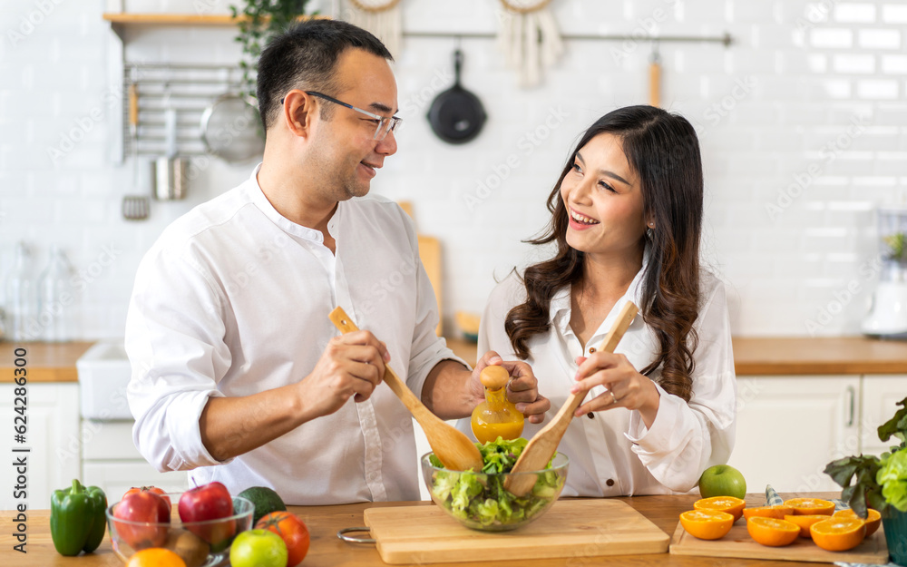 Young asian family couple having fun cooking and preparing cook vegan food healthy eat with fresh vegetable salad on counter in kitchen at home.Happy couple looking to preparing food Young asian family couple having fun cooking and preparing cook vegan food healthy eat with fresh vegetable salad on counter in kitchen at home.Happy couple looking to preparing food