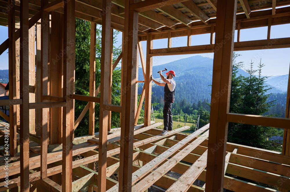 Carpenter constructing wooden frame, two-story house near forest. Bearded man hammering nails with hammer while wearing protective helmet and work coveralls. Concept of modern ecological construction.
