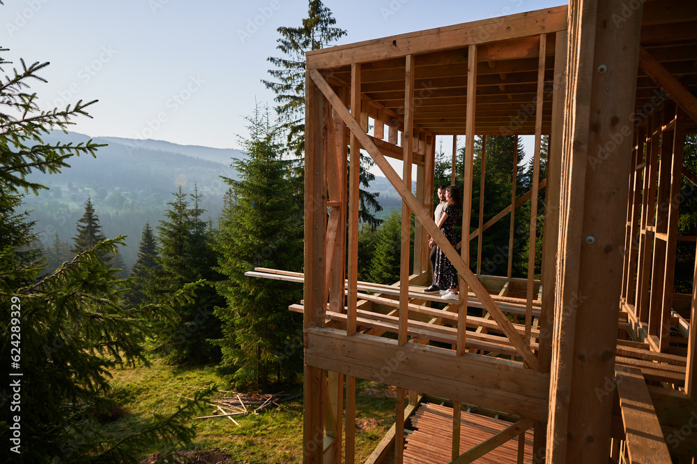 Man and woman inspecting their future wooden frame house nestled in the mountains near forest. Youthful couple at construction site in early morning. Concept of contemporary ecological construction. Man and woman inspecting their future wooden frame house nestled in the mountains near forest. Youthful couple at construction site in early morning. Concept of contemporary ecological construction.