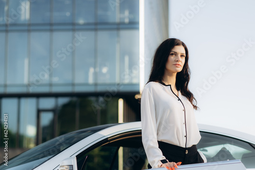 Portrait of confident stylish businesswoman holding mobile phone looking at camera standing near car, copy space. Transportation concept. © Roman