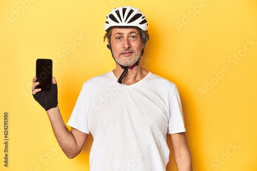Cyclist man showing phone on yellow backdrop happy, smiling and cheerful.
