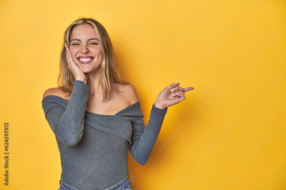 Elegant blonde on yellow backdrop, holds copy space on a palm, keep hand over cheek. Amazed and delighted. Elegant blonde on yellow backdrop, holds copy space on a palm, keep hand over cheek. Amazed and delighted.