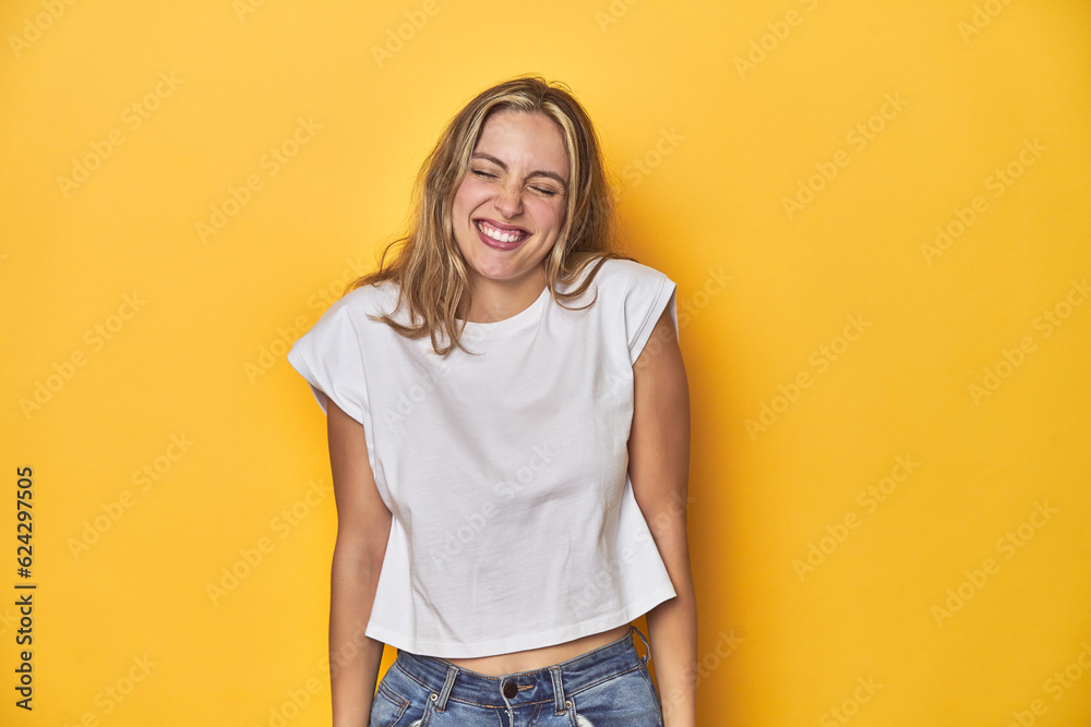 Young blonde Caucasian woman in a white t-shirt on a yellow studio background, laughs and closes eyes, feels relaxed and happy. Young blonde Caucasian woman in a white t-shirt on a yellow studio background, laughs and closes eyes, feels relaxed and happy.
