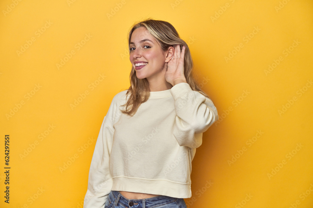 Young blonde Caucasian woman in a white sweatshirt on a yellow studio background, trying to listening a gossip. Young blonde Caucasian woman in a white sweatshirt on a yellow studio background, trying to listening a gossip.
