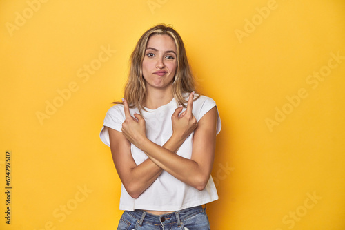 Young blonde Caucasian woman in a white t-shirt on a yellow studio background, points sideways, is trying to choose between two options.