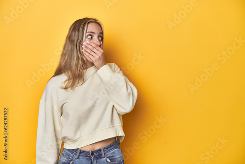 Young blonde Caucasian woman in a white sweatshirt on a yellow studio background, thoughtful looking to a copy space covering mouth with hand.