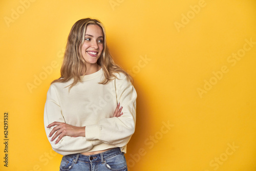 Young blonde Caucasian woman in a white sweatshirt on a yellow studio background, smiling confident with crossed arms. photo