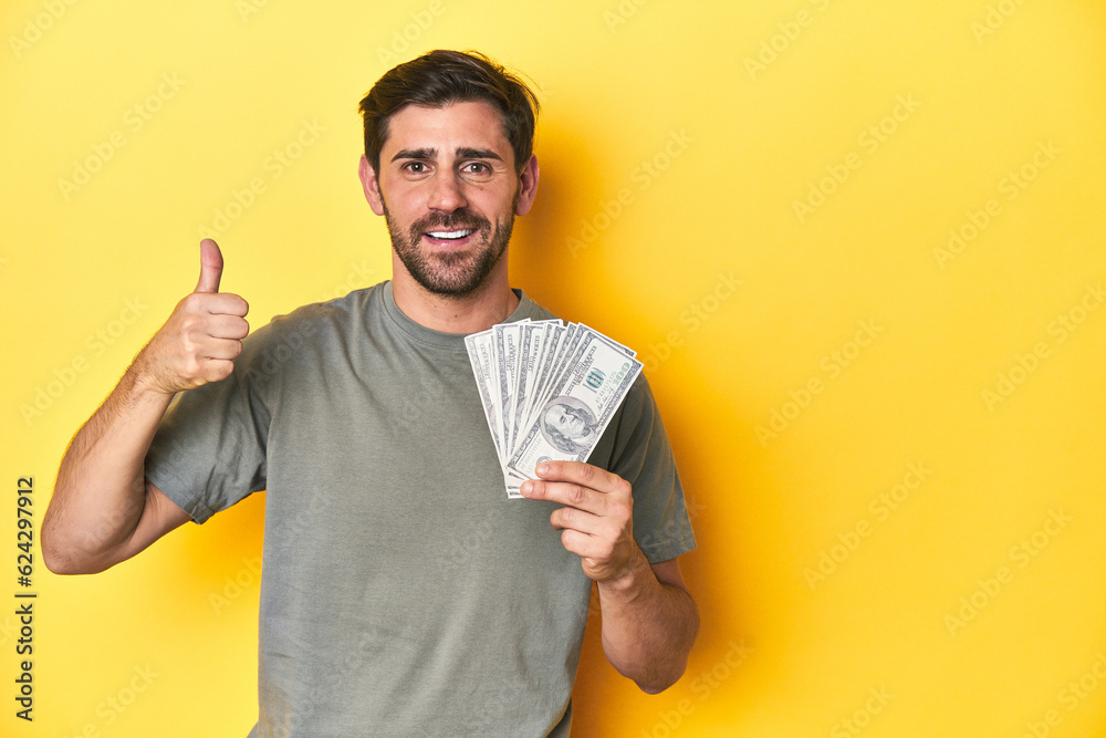 Caucasian man holding dollars, yellow studio shot smiling and raising thumb up Caucasian man holding dollars, yellow studio shot smiling and raising thumb up