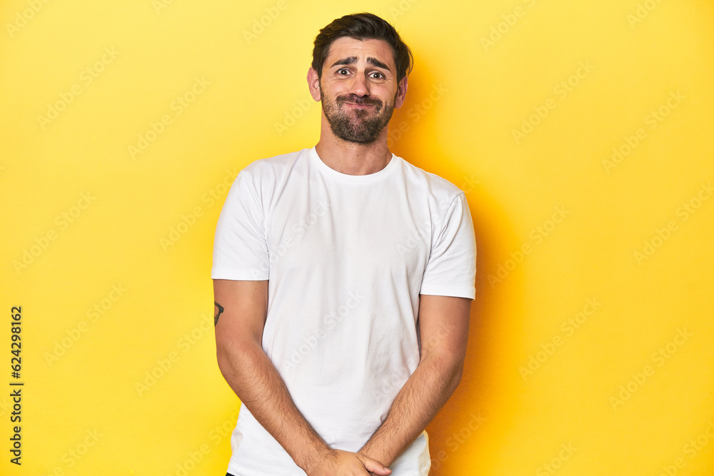 Caucasian man in white t-shirt on yellow studio background blows cheeks, has tired expression. Facial expression concept. Caucasian man in white t-shirt on yellow studio background blows cheeks, has tired expression. Facial expression concept.