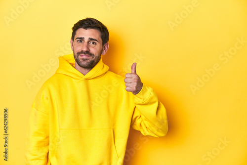 Man in yellow hoodie, monochrome studio backdrop smiling and raising thumb up