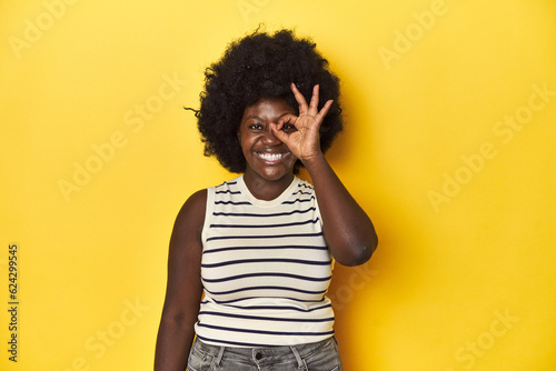 African-American woman with afro  studio yellow background excited keeping ok gesture on eye.