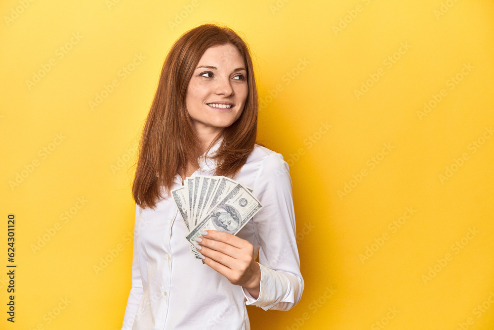 Redhead with dollar bills, cash in hand looks aside smiling, cheerful and pleasant. Redhead with dollar bills, cash in hand looks aside smiling, cheerful and pleasant.