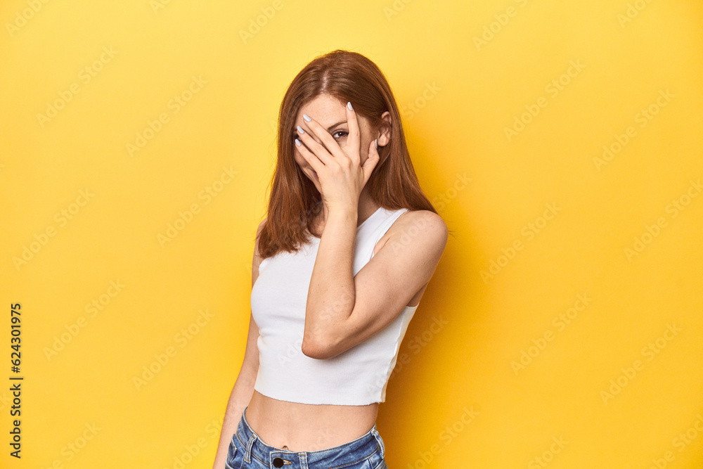 Redhead in white tank top, relaxed studio pose blink at the camera through fingers, embarrassed covering face. Redhead in white tank top, relaxed studio pose blink at the camera through fingers, embarrassed covering face.