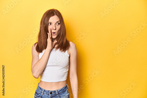 Redhead in white tank top, relaxed studio pose is saying a secret hot braking news and looking aside