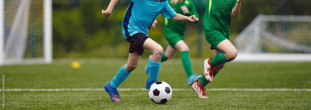 Obraz premium Junior level soccer competition between players running and kicking soccer. ball. Anonymous youth junior athletes in red and blue soccer shirts