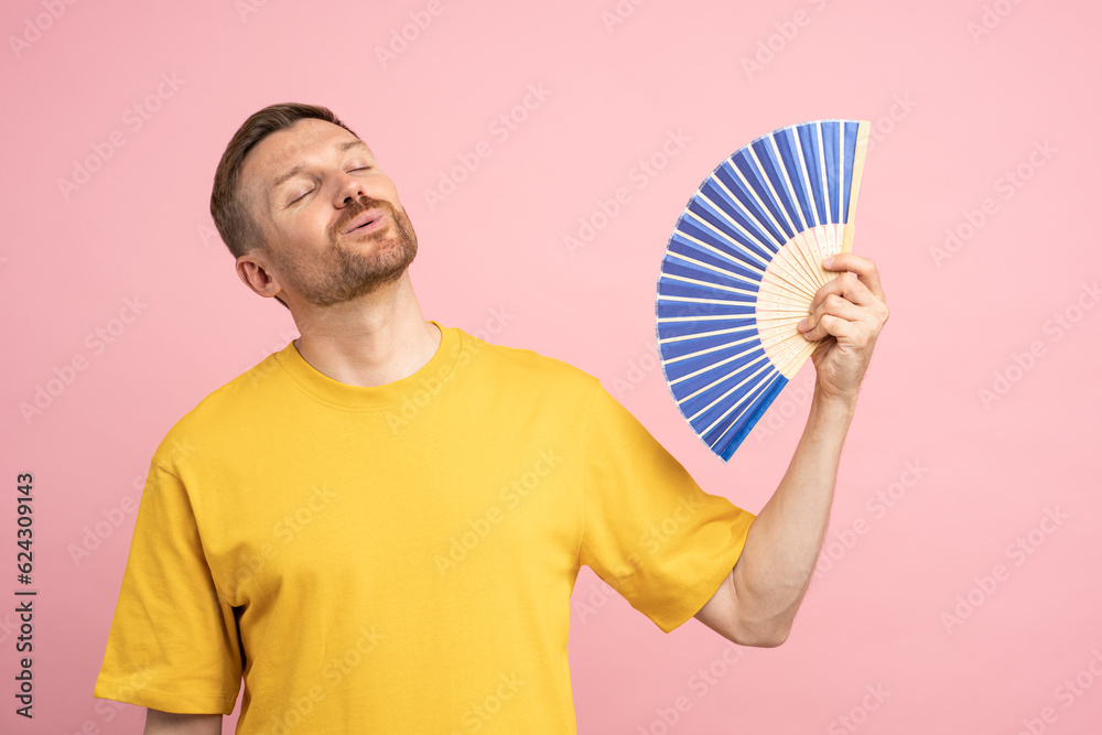 Overheated man using paper fan suffer from heat, sweating, cools herself due problem no air conditioner at home at summer weather, enjoying fresh air and closed eyes isolated on studio pink background Overheated man using paper fan suffer from heat, sweating, cools herself due problem no air conditioner at home at summer weather, enjoying fresh air and closed eyes isolated on studio pink background