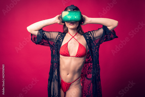 Studio photo shoot of beautiful brown hair model posing wearing red swimwear and vr glasses with red background behind. Sexy woman wearing futuristic augmented reality goggles