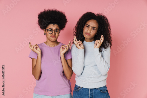 Two displeased african women holding fingers crossed for good luck isolated over pink background