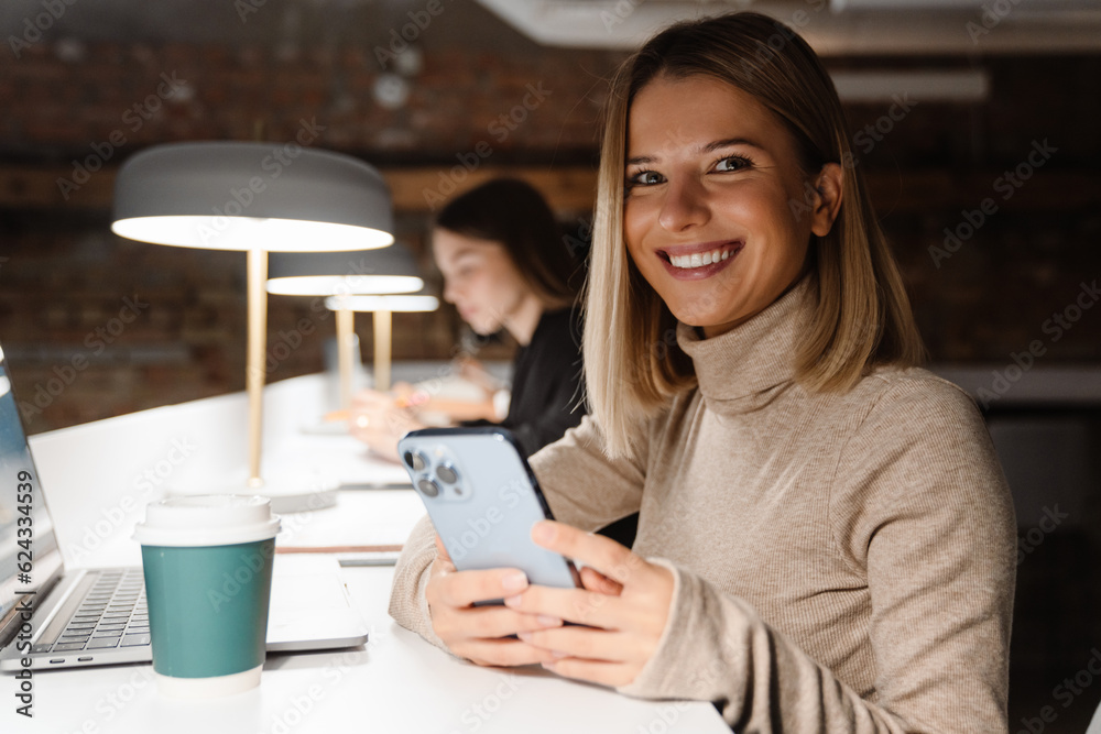 Smiling woman using mobile phone while sitting at table with her colleague on the background Smiling woman using mobile phone while sitting at table with her colleague on the background