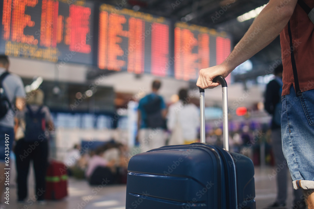 Traveling by airplane. Man waiting in airport terminal. Selective focus on hand holding suitcase against arrival and departure board. Passenger is ready for travel..