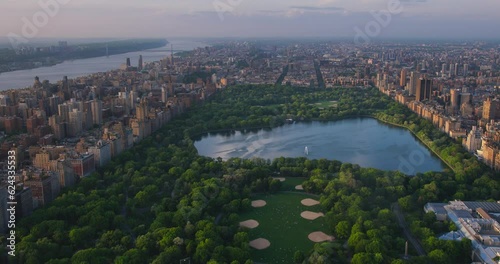 Aerial Helicopter Footage Over Central Park with Nature, Trees, People Having Picnic and Resting on a Field Around Manhattan Skyscrapers Cityscape. Beautiful Evening with Warm Sunset Light