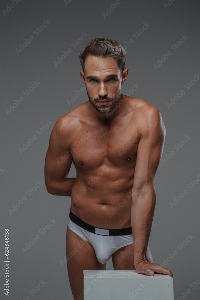 An attractive male model with a handsome physique and bare torso, posing in underwear while leaning on a gray crate, set against a gray background