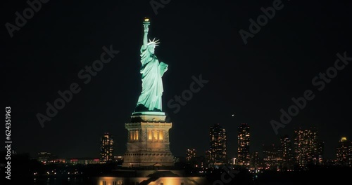 Aerial Helicopter Cinematic View of the Statue of Liberty with Manhattan Skyline Cityscape with Lights in Office Buildings and Skyscrapers at Night. Panoramic View of New York City Skyline