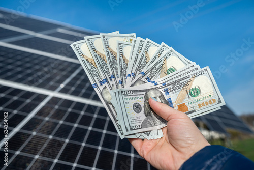 with dollar bill in his hand shows solar panels at station which reflect sun's rays.