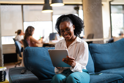 Beautiful African businesswoman sitting on sofa in office. Young woman using digital tablet..