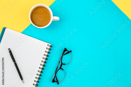 Notebook, eye glasses and cup of coffee on a blue and yellow background.