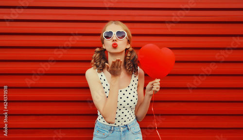 Portrait of happy cute girl with big red heart shaped balloon and girly hairstyle blows her lips sends air kiss wearing sunglasses on background