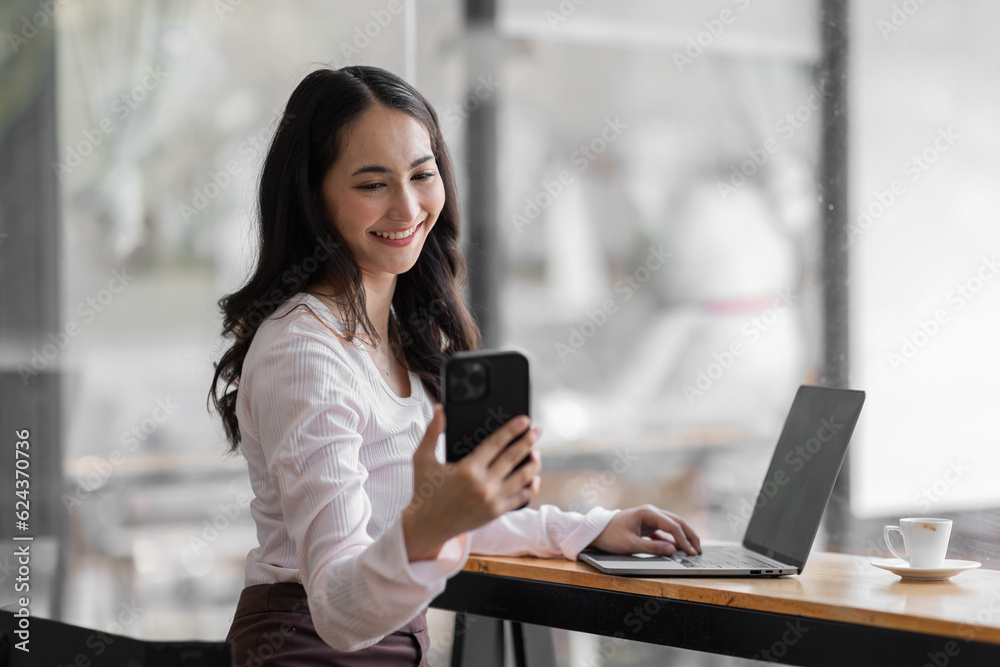 Fototapeta premium Asian Woman entrepreneur busy with her work in the office. Young Asian woman using phone or cellphone while working on computer at her desk.