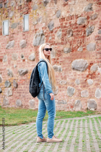 Girl in sunglasses with a backpack in jeans clothes on the background of a brick building. © 7707601