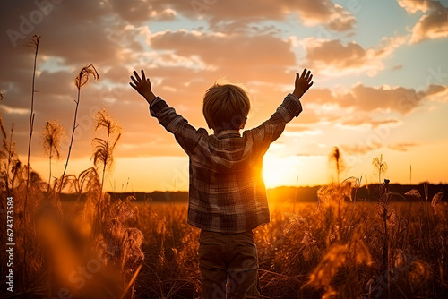 A little boy raises his hands above the sunset sky  enjoying life and nature. Happy kid on a summer field looking at the sun. Silhouette of a male child in the sun. Fresh air  environment concept.