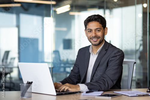 Photo Portrait of young arab businessman, man smiling and looking at camera while sitting inside office, boss in business suit at workplace using laptop