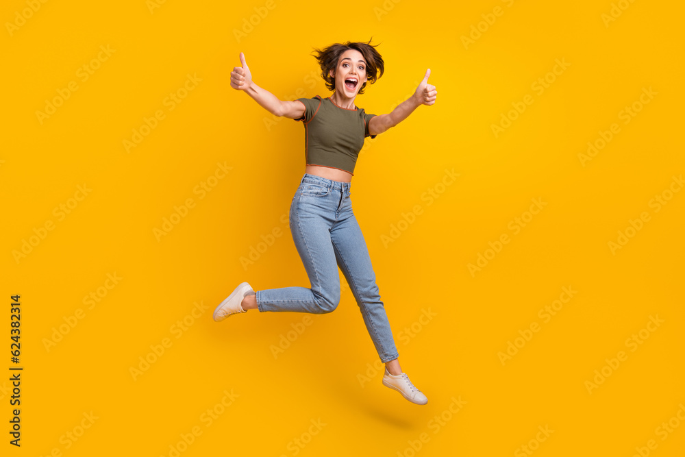 Full size photo of optimistic overjoyed girl dressed jeans khaki top jump show thumbs up approve isolated on yellow color background Full size photo of optimistic overjoyed girl dressed jeans khaki top jump show thumbs up approve isolated on yellow color background