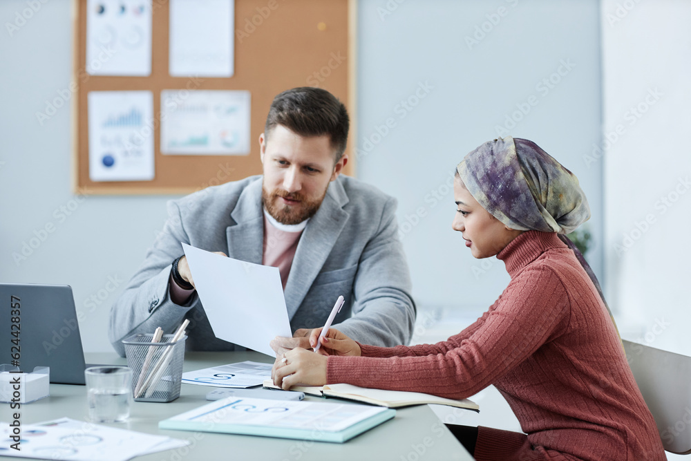 Young confident male economist explaining data in financial document to female colleague while discussing working points at meeting in office Young confident male economist explaining data in financial document to female colleague while discussing working points at meeting in office