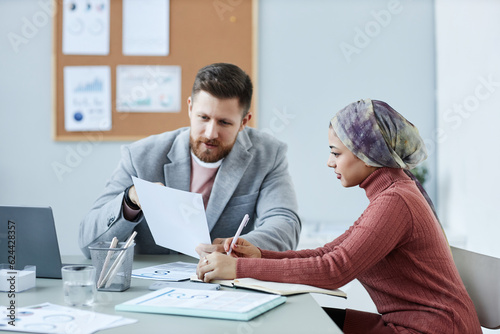 Young confident male economist explaining data in financial document to female colleague while discussing working points at meeting in office